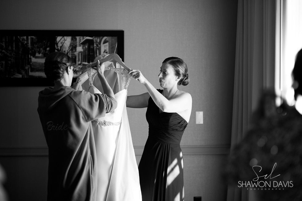 Two people adjusting a white dress on a hanger indoors near a window