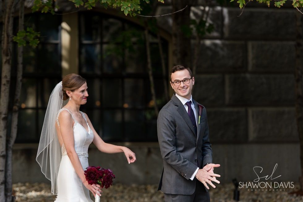 Bride in white dress and groom in gray suit smiling outdoors, holding hands during wedding ceremony