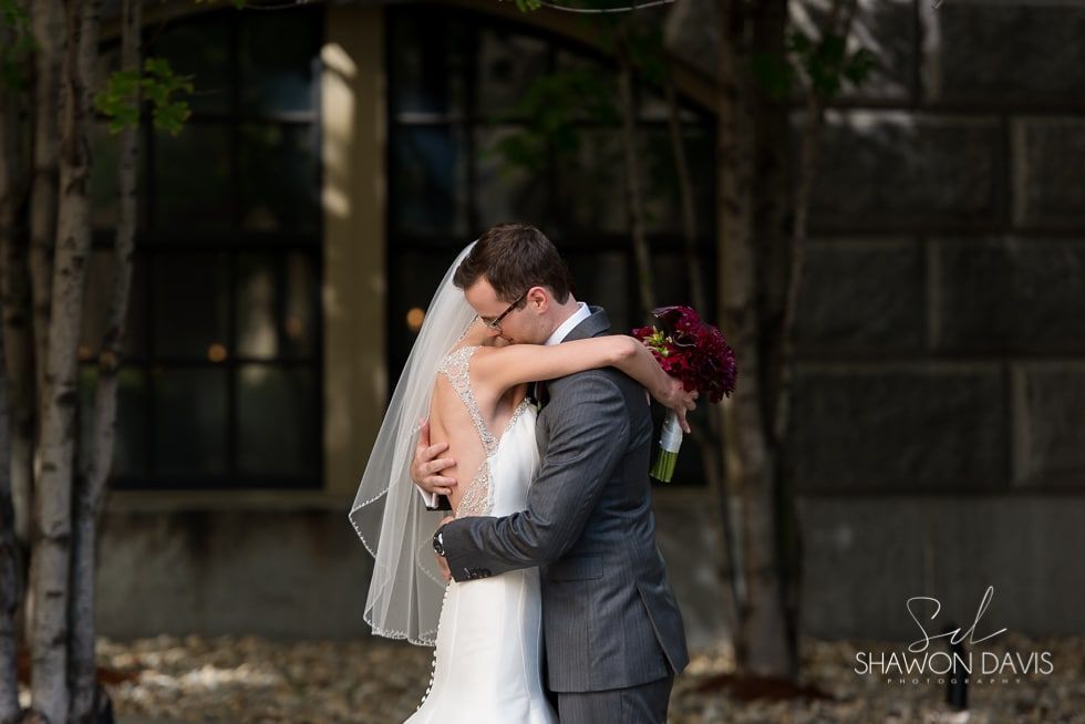 Bride and groom embracing outdoors, with veil and bouquet against a dark stone wall