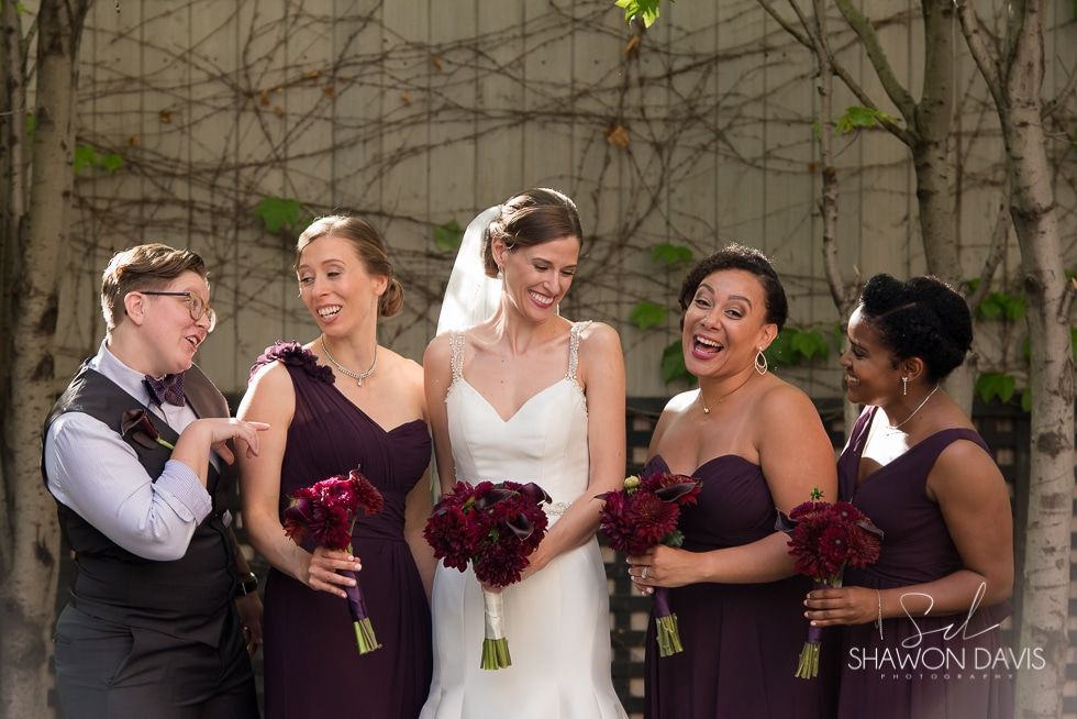Five smiling wedding party members hold purple bouquets outdoors at a wedding venue.