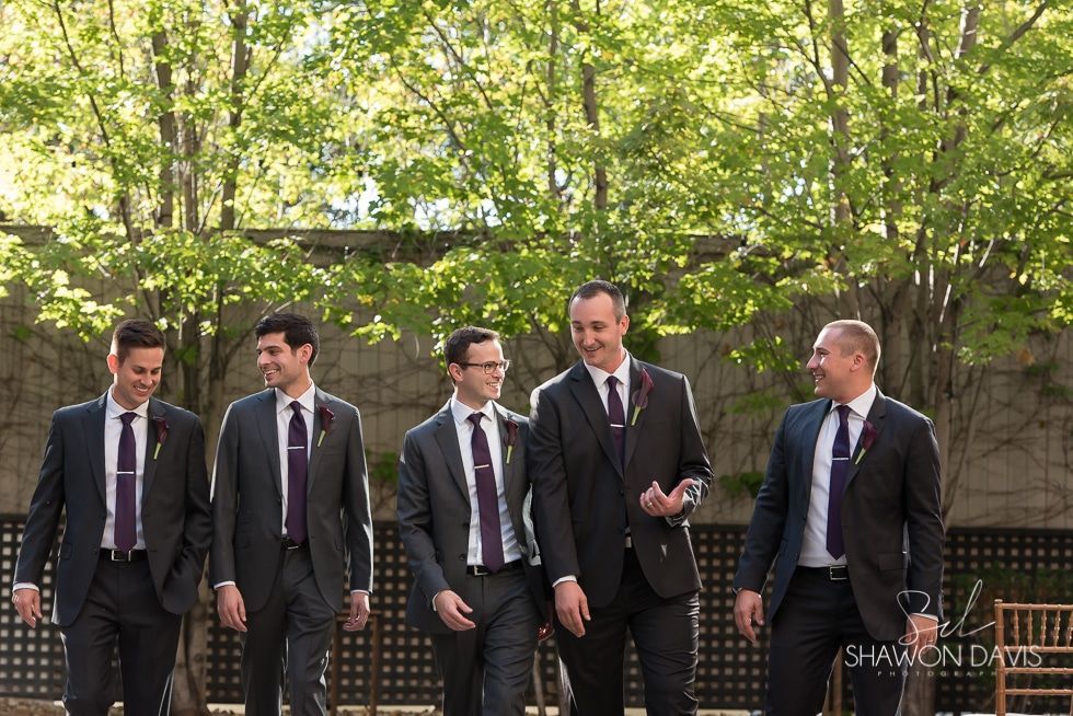 Five men in suits walking outdoors and talking in a wooded area.