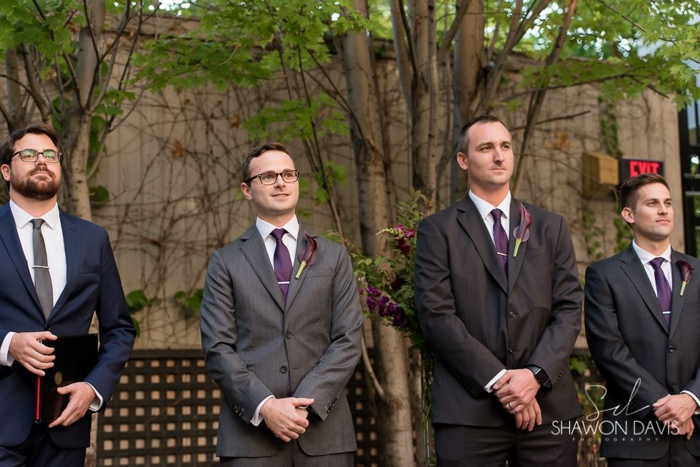Four men in suits stand outdoors during a formal event, with greenery and stone walls behind them.
