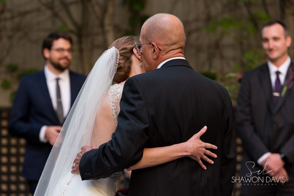 Bride in white veil embracing an older man in a suit during an outdoor wedding ceremony
