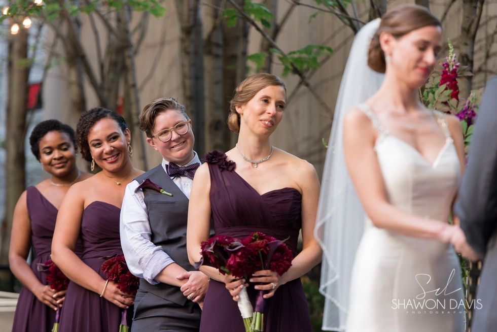 Bridesmaids in purple dresses and a white-shirted groomsman stand in line at an outdoor wedding ceremony