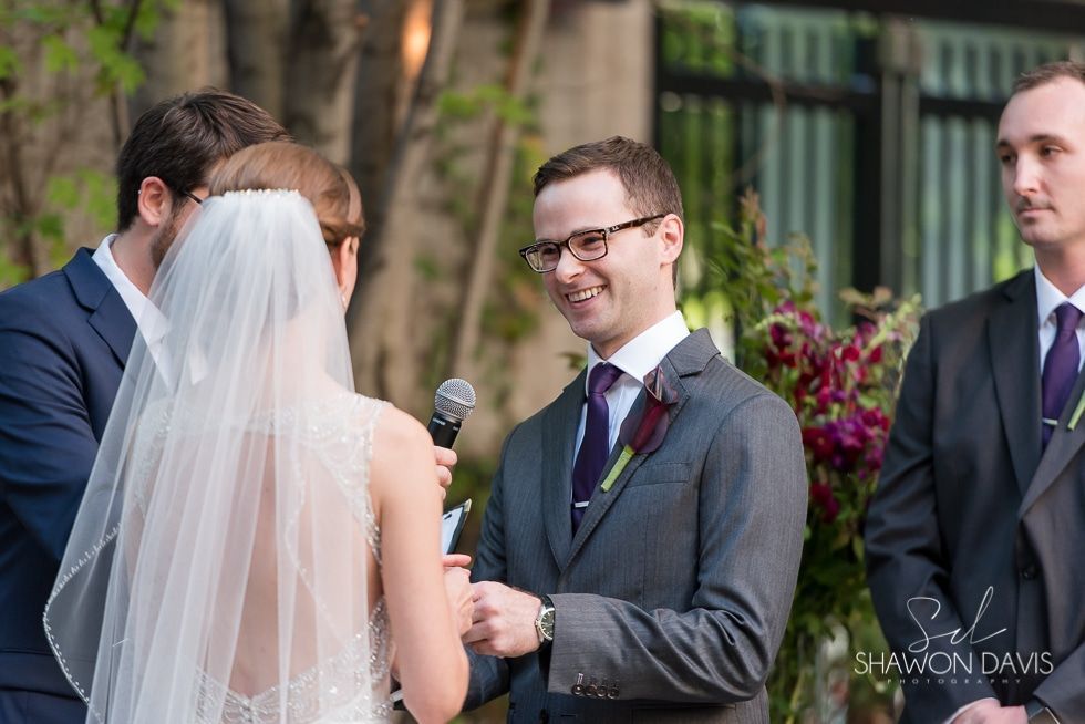 Bride in white veil faces groom holding a microphone during an outdoor wedding ceremony
