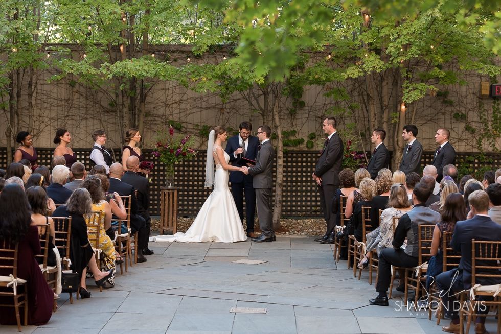 Outdoor wedding ceremony with bride and groom exchanging vows in a garden courtyard, guests seated on both sides.