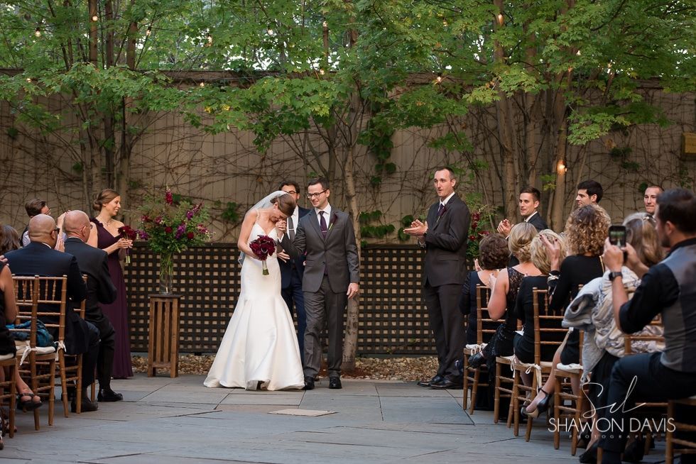 Wedding ceremony outdoors with bride, groom, officiant, and seated guests in a garden courtyard