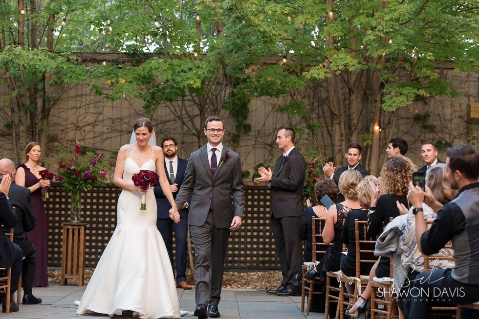 Wedding couple walking down an outdoor aisle with guests applauding under trees