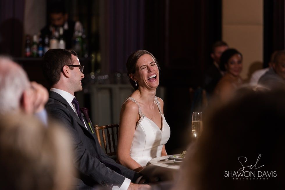 Laughing bride in white dress and groom in suit seated at a warmly lit wedding reception