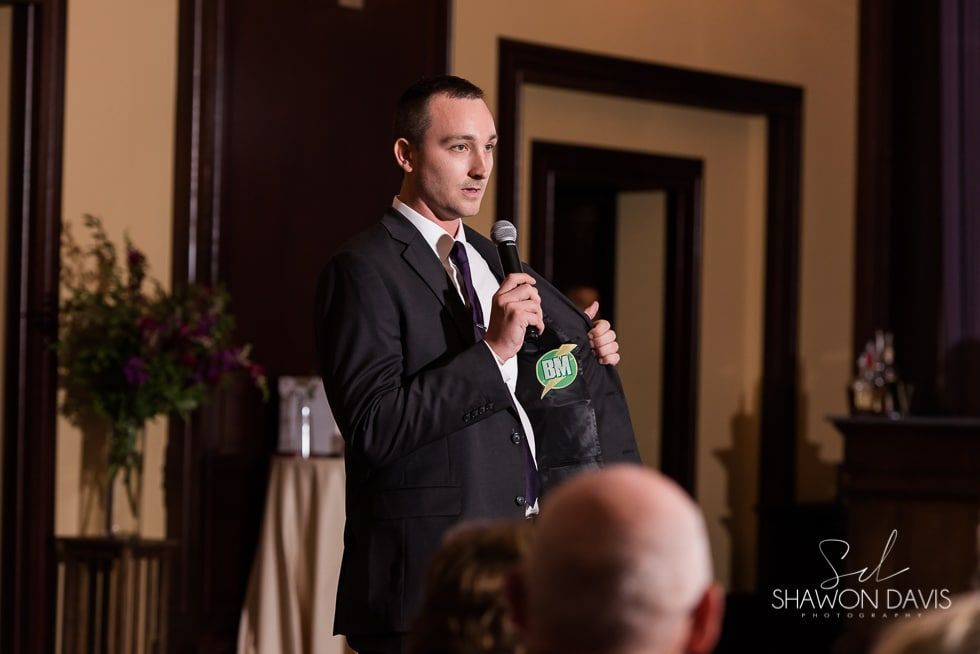 Man speaking into a microphone at a formal indoor event, wearing a dark suit with a green ribbon badge.