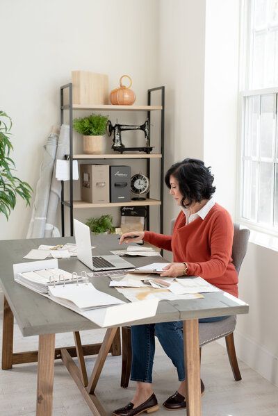 Woman working on a laptop at a dining table in a bright home office, surrounded by papers and shelves.