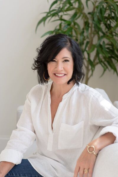Woman in a white blouse seated on a white sofa, smiling with a plant in the background.
