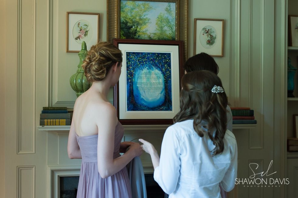 Two women view a framed blue abstract painting on a mantel in a bright room.