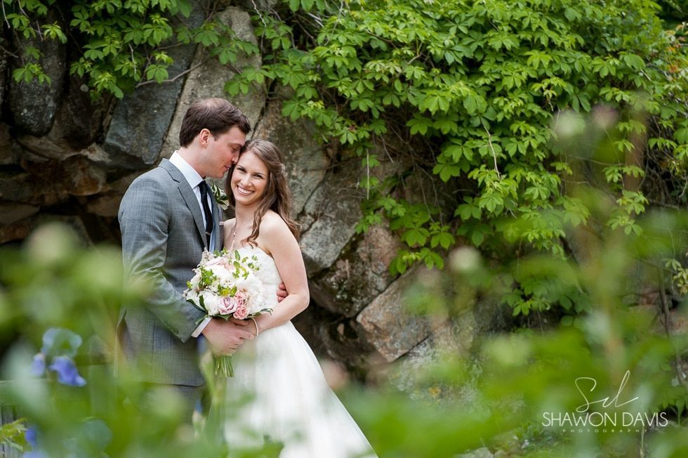 Bride and groom embracing in a garden beside rocks, with green foliage and flowers in the foreground