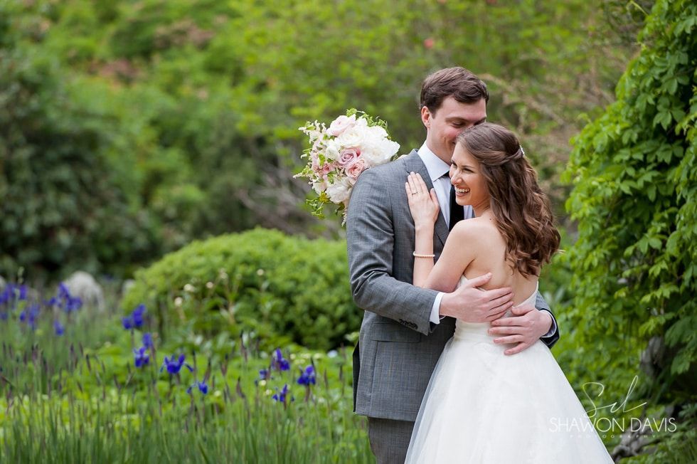 Couple embracing in a green garden, bride holding bouquet and groom in gray suit.
