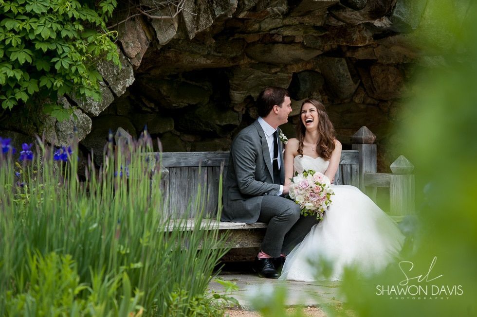 Bride and groom sitting on a bench in a garden, smiling beneath a rocky archway, with purple flowers in the foreground
