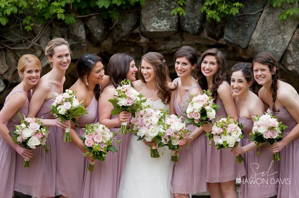 Bridesmaids in pink dresses holding bouquets, smiling outdoors in front of a stone wall.