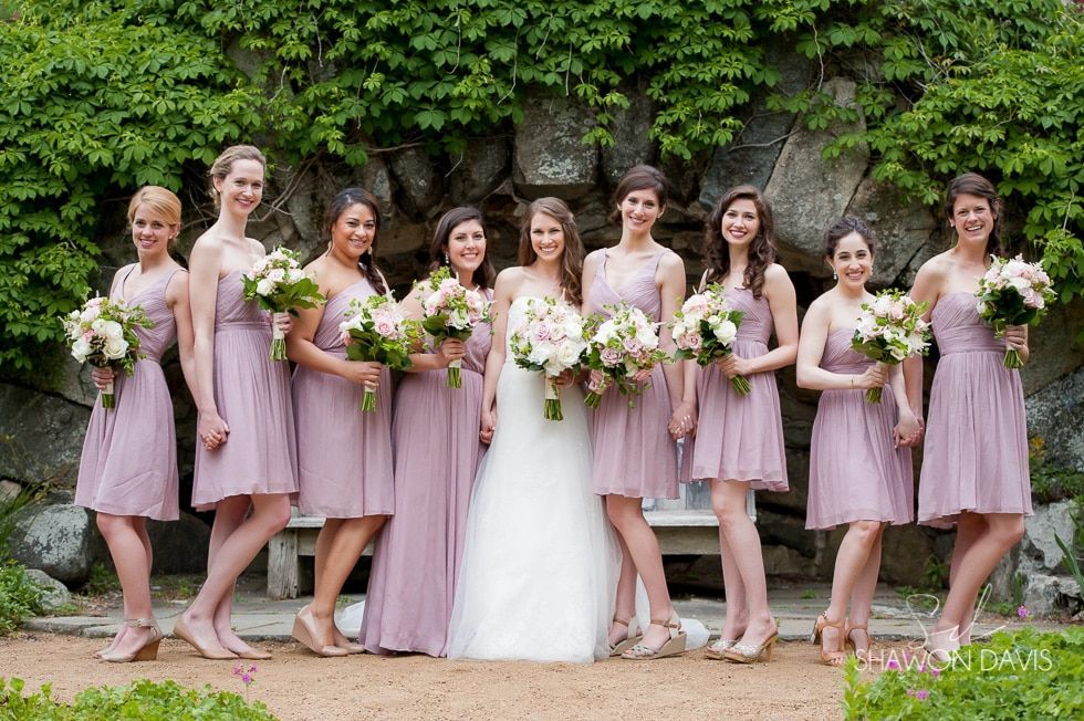 Wedding party posing outdoors, bridesmaids in blush dresses holding bouquets beside the bride in white.
