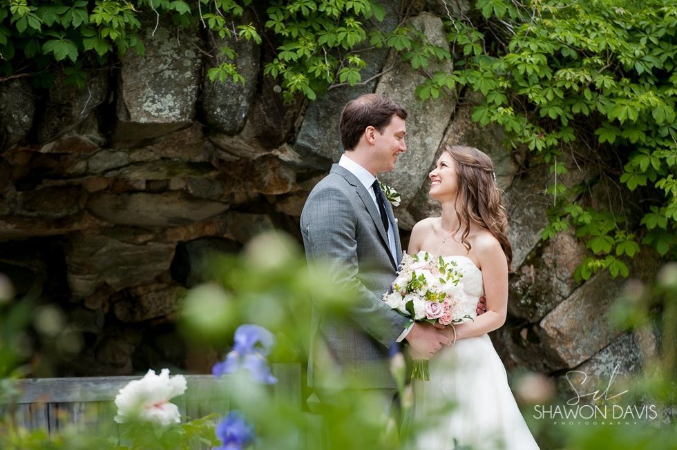 Bride and groom smiling in a leafy garden, holding a bouquet beside a stone wall