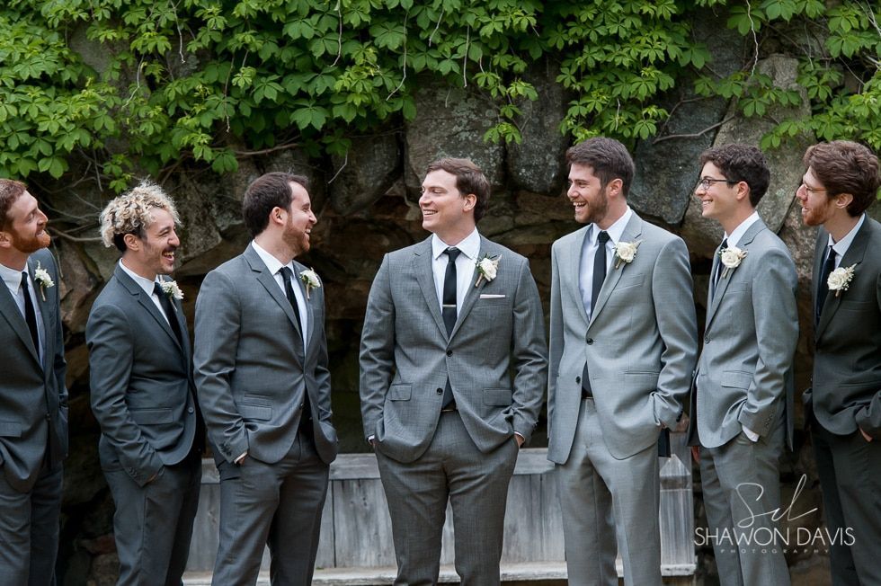 Six men in gray suits standing outdoors, smiling and talking in front of green foliage.