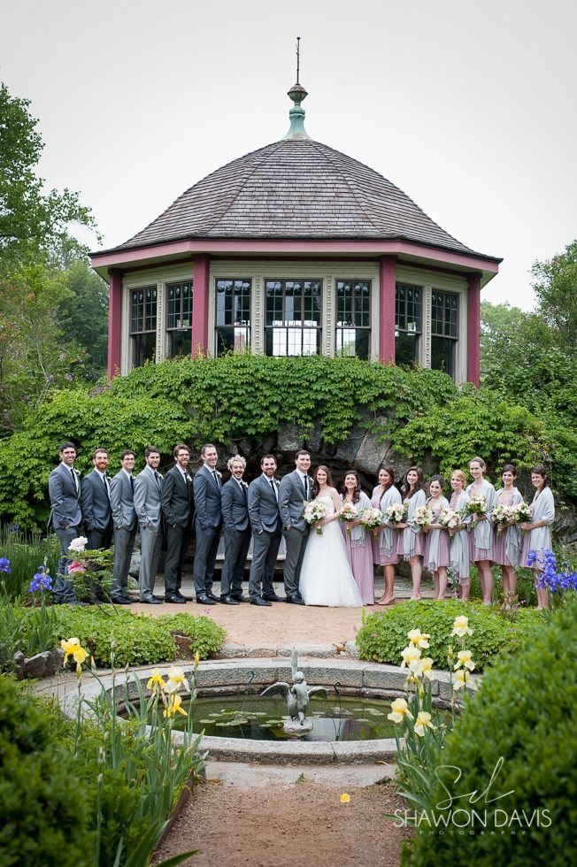 Wedding party posed in front of a pink gazebo, with a fountain and garden flowers in the foreground
