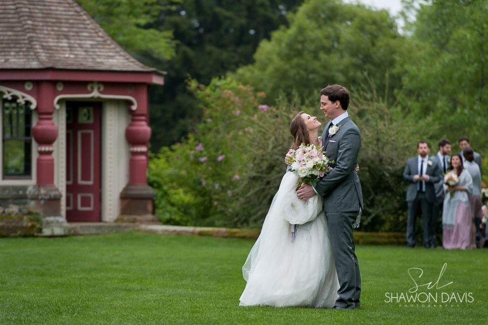 Bride and groom embracing on a lawn near a gazebo, with wedding guests in the background.