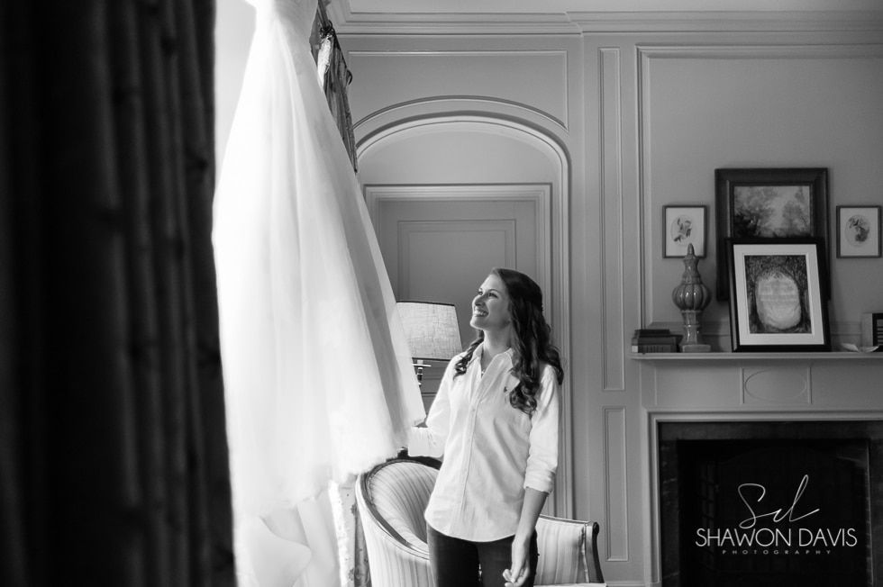 Woman standing by a window in a bright room, looking out beside a lamp and framed photos.