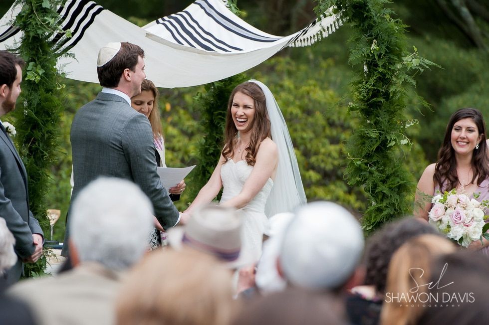 Bride smiling at outdoor wedding ceremony, holding hands with groom beneath white drapery.