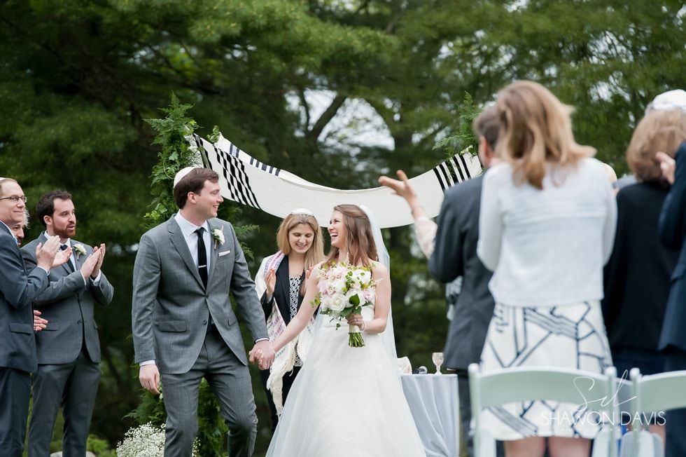 Bride and groom walking hand in hand at an outdoor wedding ceremony, guests applauding nearby.
