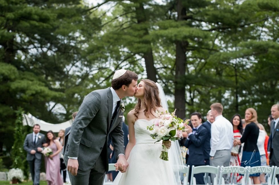 Bride and groom kiss at an outdoor wedding ceremony with guests and trees in the background