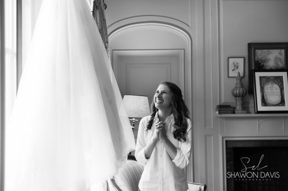 Woman in white dress standing by a bright window, smiling in a softly lit room with framed art.