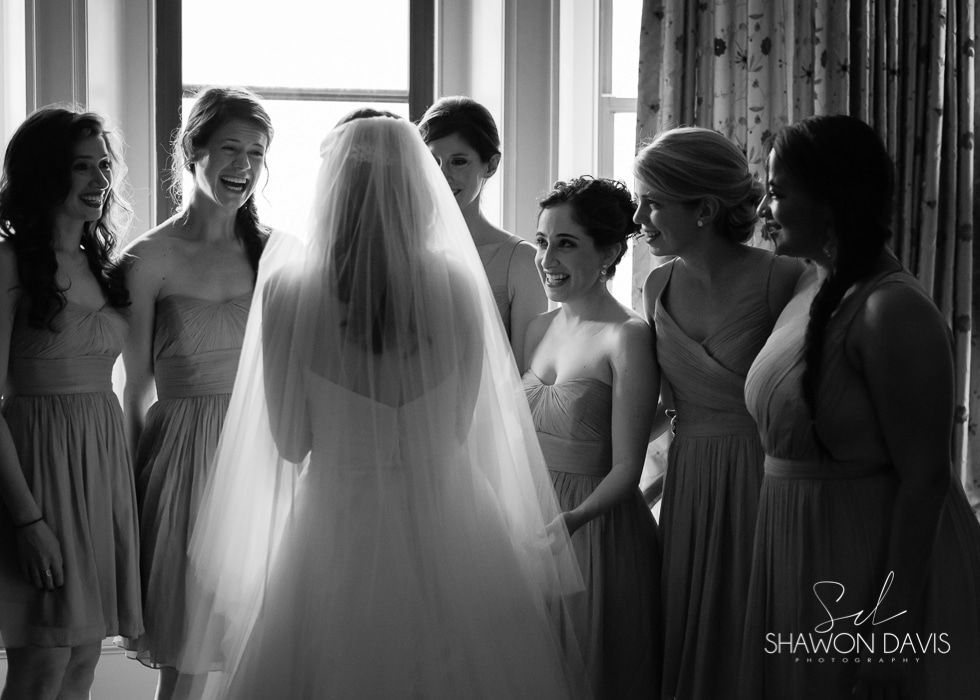 Bride with veil surrounded by bridesmaids in a sunlit room, smiling and looking at her.