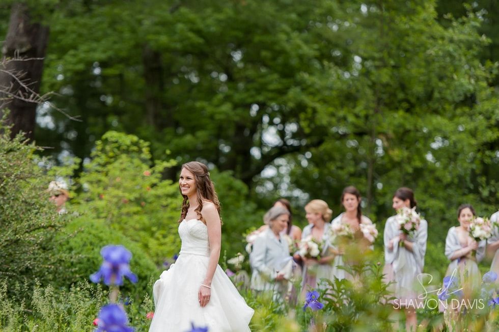 Bride in a white gown walks through a green garden with bridesmaids holding bouquets behind her.