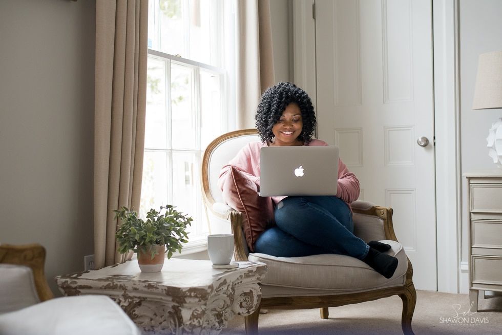 Person using a laptop on a chaise in a bright living room with curtains and potted plants