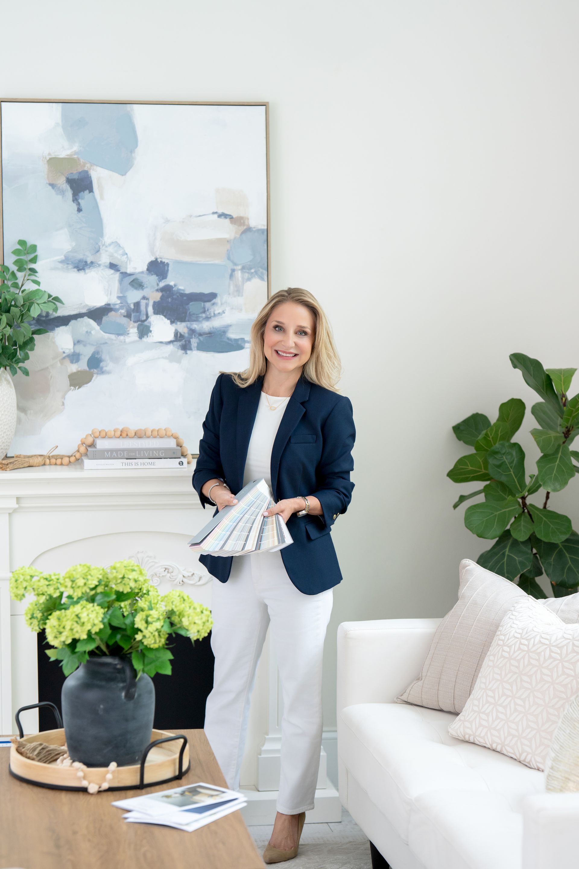 Woman seated on a beige sofa in a bright living room with plants and shelving.