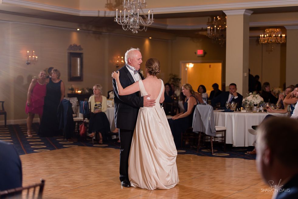 Couple slow-dancing in a ballroom at a formal event, guests seated around the dance floor.