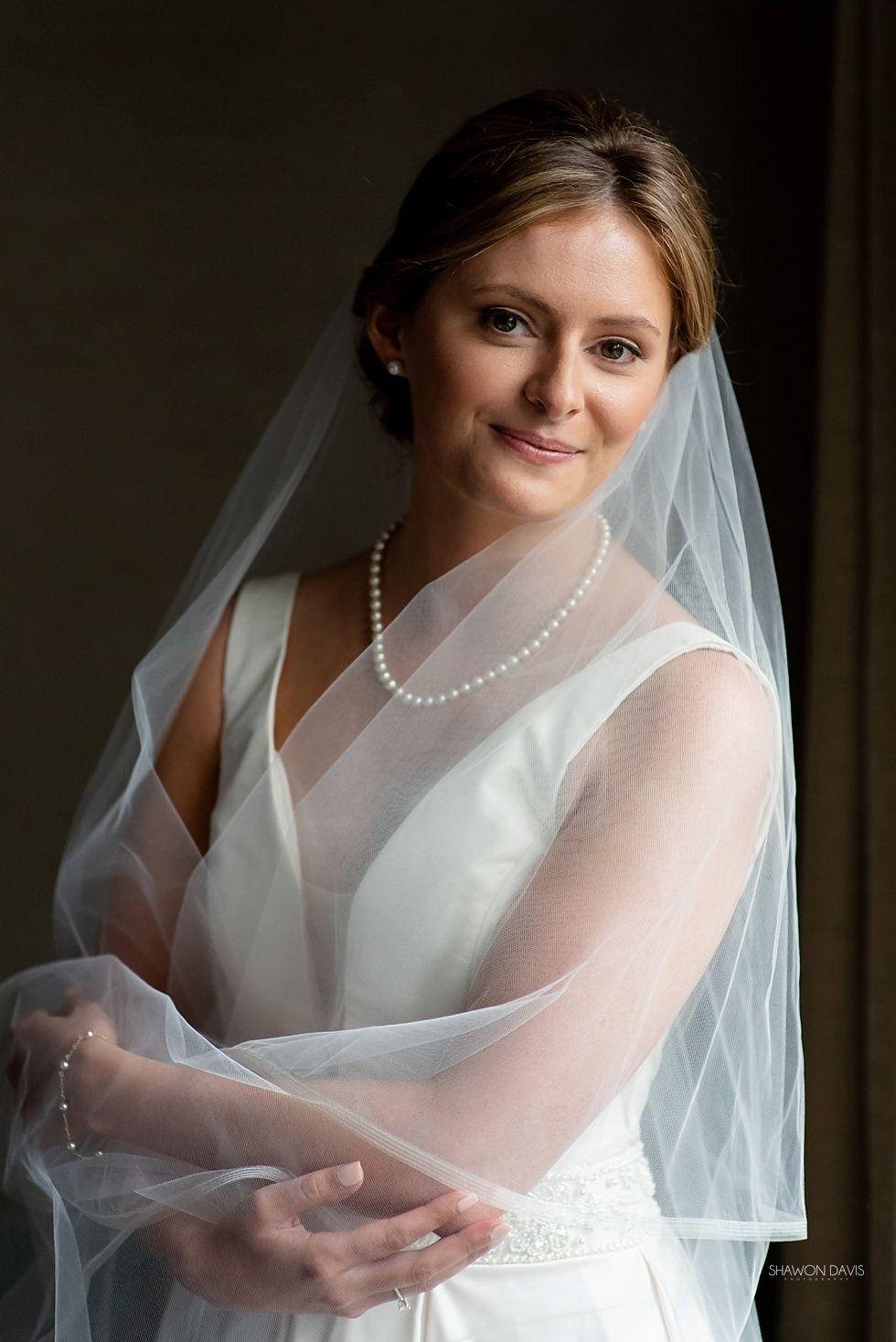 Bride in white veil and dress, smiling softly with hands clasped against a dark background