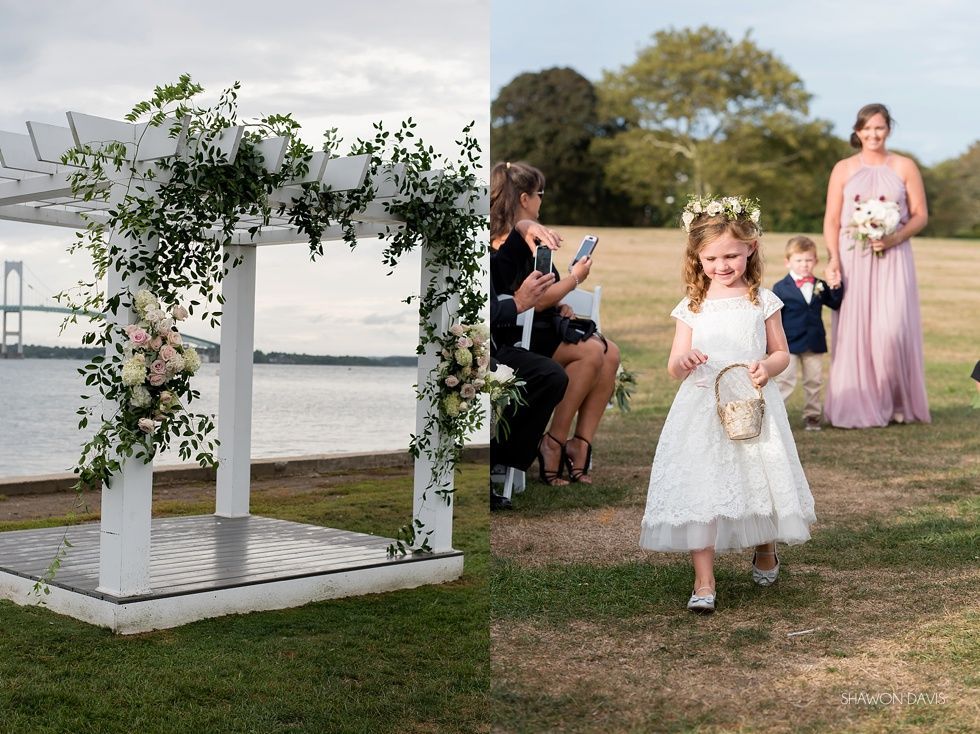 Flower girl in white dress walks past a floral arch at an outdoor wedding by the water