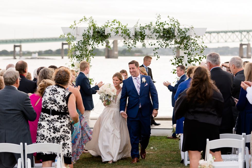 Wedding couple walking down aisle outdoors by a river, guests applauding under a floral arch.