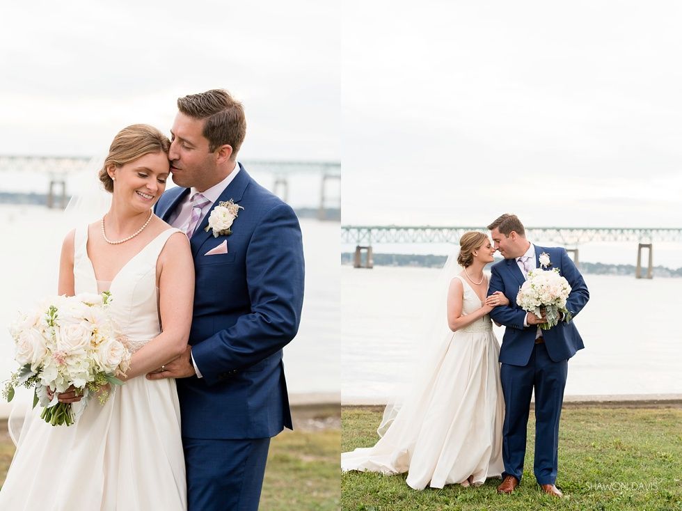 Bride and groom embracing by the water with a bridge in the background
