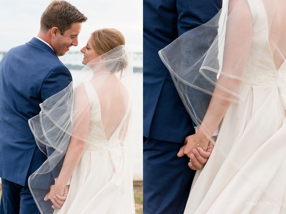 Bride and groom holding hands, smiling under a flowing veil by the water