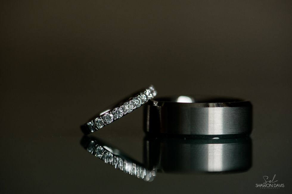 Diamond engagement ring leaning against black wedding band on reflective dark surface
