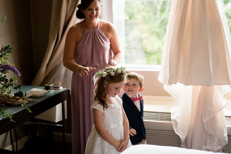 Young flower girl and ring bearer by a window as a woman adjusts the girl’s floral crown