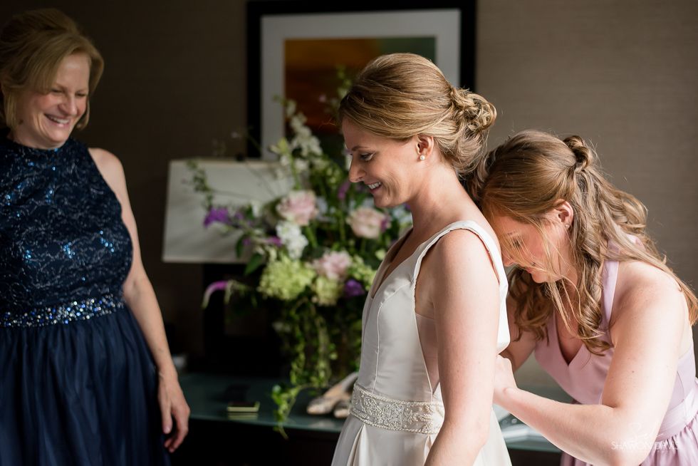 Three women in formal dresses adjusting a bride’s gown indoors