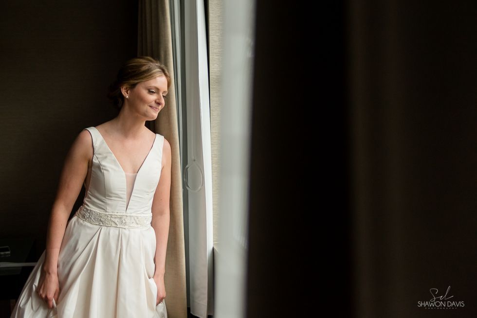 Woman in a white dress standing by a window, looking outside in soft light.