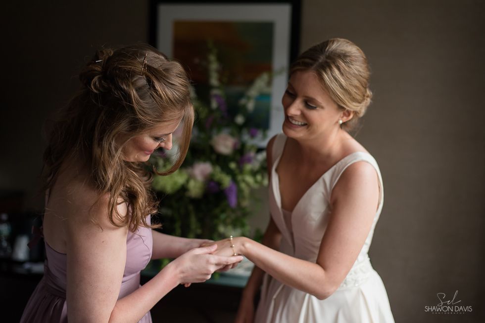 Two women smiling as one places a ring on the other's finger indoors during a ceremony
