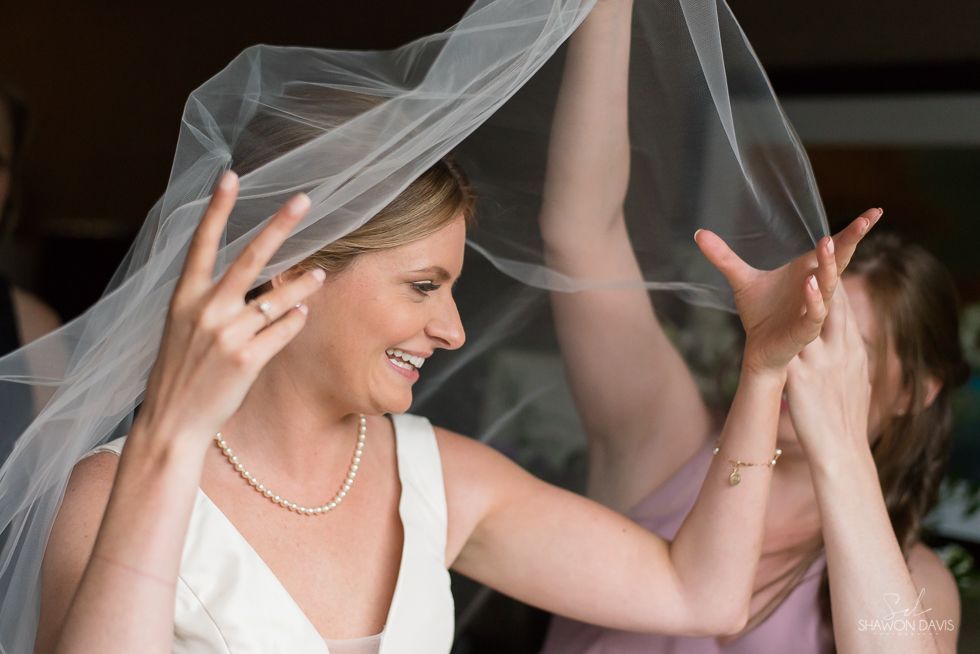 Bride smiling as women lift a sheer veil during a wedding celebration