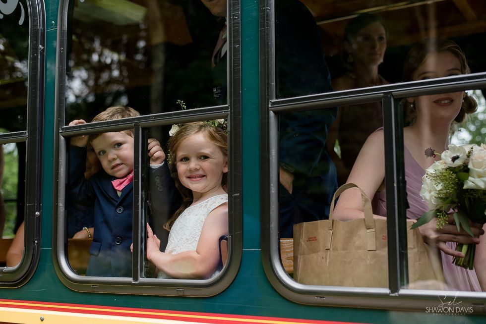 Children and a woman ride in a vintage green bus, smiling and holding flowers
