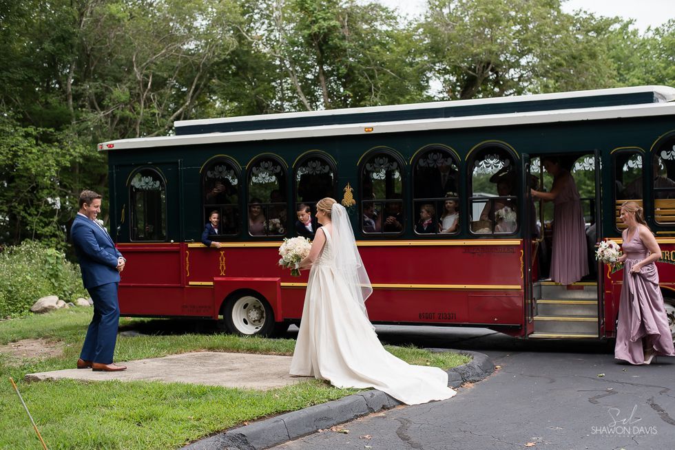 Bride in white dress beside a red trolley, with guests boarding in a green park setting