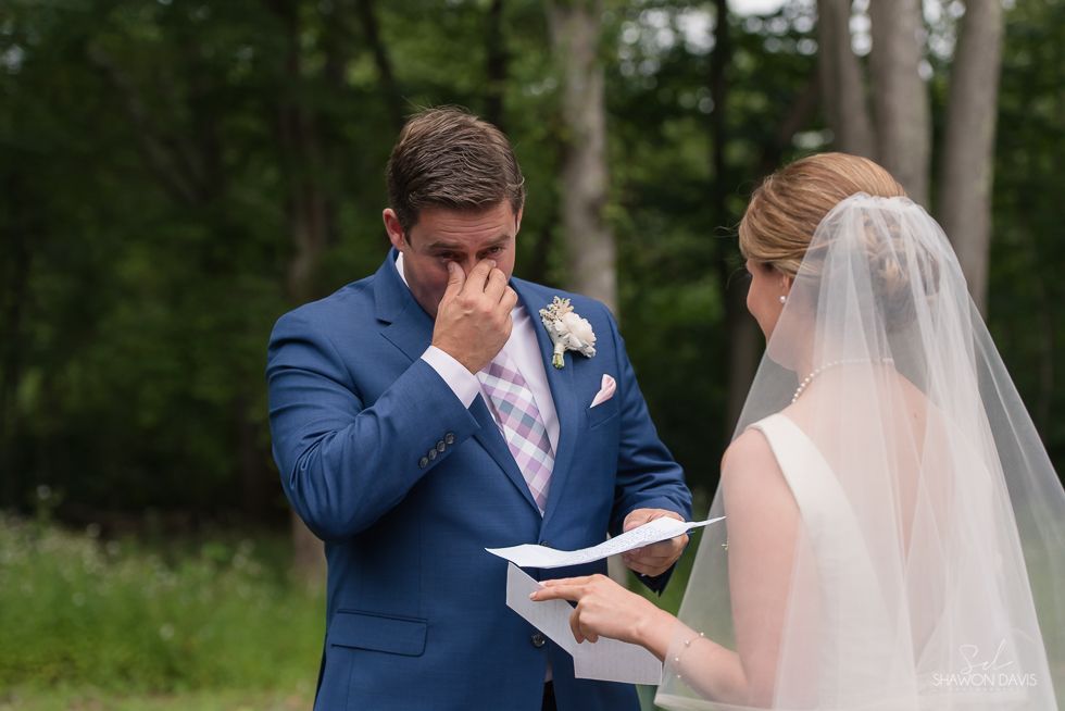 Bride and groom exchanging vows outdoors; groom wipes tears as bride reads from a paper.